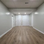 Spacious empty basement with wooden flooring and white doors.